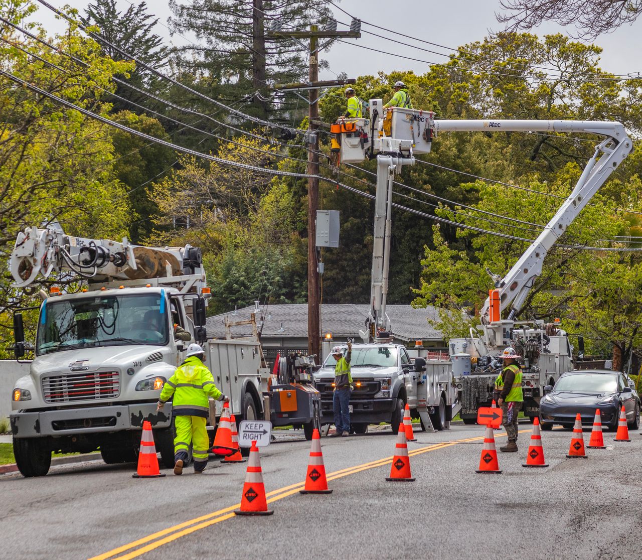 heros-img Utility workers in reflective gear repair power lines using bucket trucks and cones on a suburban street.