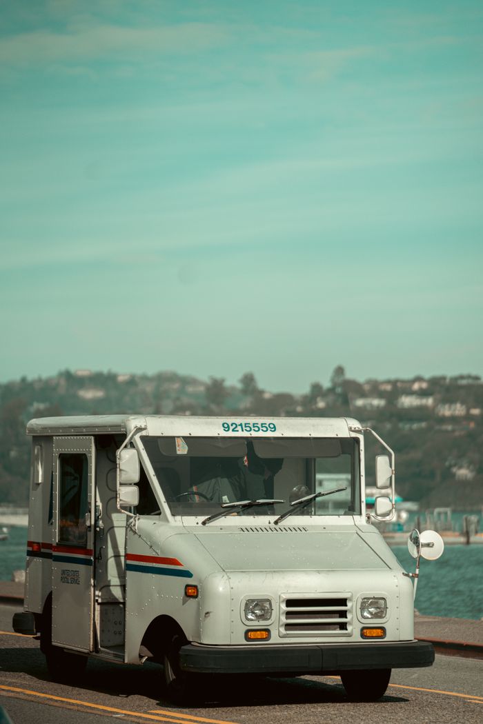who-we-are Postal delivery truck on a coastal road in Sausalito, CA, with a scenic backdrop.